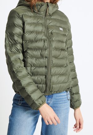 Kvinde iført olivengrøn quiltet dunjakke med lynlås og hætte, kombineret med lyseblå denimjeans, stående mod en hvid baggrund.