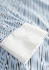 Közeli felvétel egy kék és fehér csíkos ing ujja, amelynek fehér, gombos mandzsettája van a tetején.