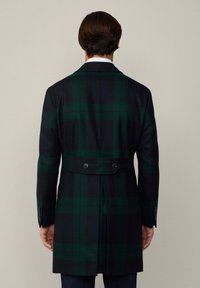 Kabát má tmavozelený a námornícky pásikový vzor, strih na mieru, dvojité gombíky v páse a vetvený chrbát pre väčšiu pohyblivosť.