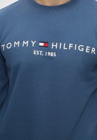 Μπλε φούτερ με κεντημένα λευκά γράμματα "Tommy Hilfiger", ένα μικρό κόκκινο, λευκό και μπλε λογότυπο, και "EST. 1985" από κάτω.