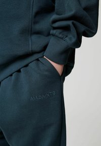 Tamsiai mėlynas džemperis ir kelnės, pagamintos iš tekstūruotos medžiagos, su matomu siūlu ir kišene. Ant kelnių išsiuvinėtas "ALLSAINTS" logotipas.