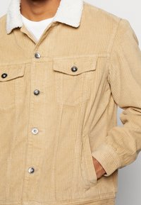 Beige manchesterjacka med vit fårskinnskrage, tryckknappar, två bröstfickor och ribbade muddar. Texturerat tyg med vertikala ränder.