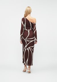 Žena s dlouhými blond vlasy má na sobě hnědé šaty s dlouhým rukávem, bílé abstraktní vzory a střihem nad rameny, které doplňuje bílými sandály na vysokém podpatku.