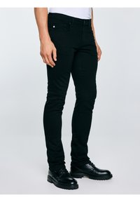 Férfi fekete, szűk szabású nadrágban és fekete bőr fűzős cipőben, világos egyszínű háttér előtt állva.