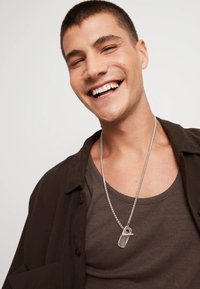 Un jeune homme souriant, portant un débardeur marron et une chemise sombre ouverte, avec un collier en chaîne argentée arborant un pendentif distinct.