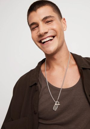 Joven sonriendo, vistiendo una camiseta sin mangas marrón y una camisa oscura abierta, con un collar de cadena plateada que presenta un colgante distintivo.