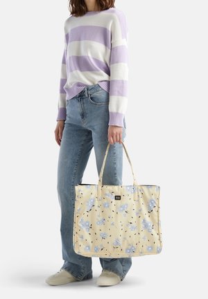 Vrouw in een lichtpaars-wit gestreepte trui en blauwe jeans, met een grote crèmekleurige draagtas met blauw bloemmotief en twee handvatten.