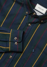 Námornícka košeľa na gombíky s vertikálnymi pruhmi v horčicovej, tmavozelenej a burgundskej farbe, s gombíkovým golierom a manžetami a vyšitým logom na hrudi.