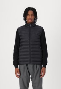 Black puffer vest with a high collar, zip closure, and horizontal quilted design, paired with a black long-sleeve top and gray trousers.