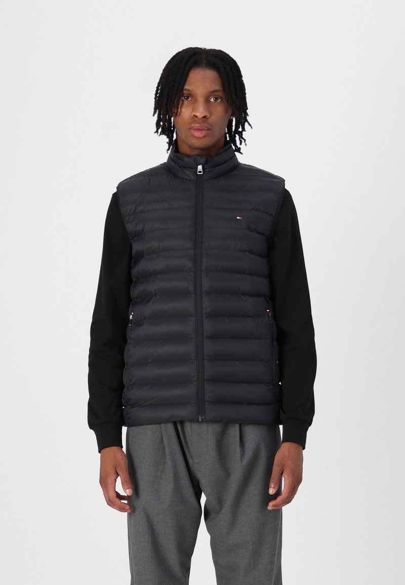 Black puffer vest with a high collar, zip closure, and horizontal quilted design, paired with a black long-sleeve top and gray trousers.