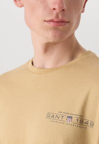 Beige bomullströja med rund halsringning och korta ärmar. Har en tryckt logotyp i marinblå och röd med textdetaljer på framsidan.