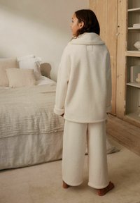 Frau steht barfuß in einer gemütlichen cremefarbenen Fleecejacke und weiten Hosen in einem Schlafzimmer mit neutraler Bettwäsche und Holzböden.