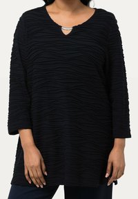Svart topp med texturerat vågmönster, V-formad halsringning prydd med silverdetaljer och trekvartslånga ärmar. Mjuk tygkänsla.
