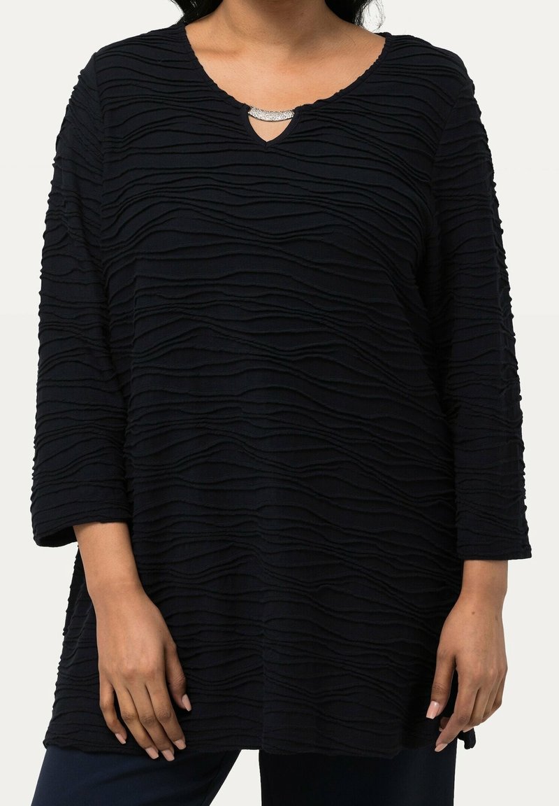 Svart topp med texturerat vågmönster, V-formad halsringning prydd med silverdetaljer och trekvartslånga ärmar. Mjuk tygkänsla.