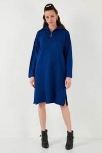 Žena v modrých šatách po kolená so zipsovým golierom stojaca pred bielym pozadím, doplnená čiernymi členkovými čižmami.