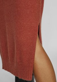 Pull tricoté couleur rouille avec ourlet côtelé et fente latérale, mettant en valeur une texture douce et une coupe ajustée le long du corps.