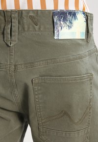 Olivové kalhoty se zakřiveným šitím zadní kapsy a látkovou nášivkou s plážovou scénou připevněnou na pase.