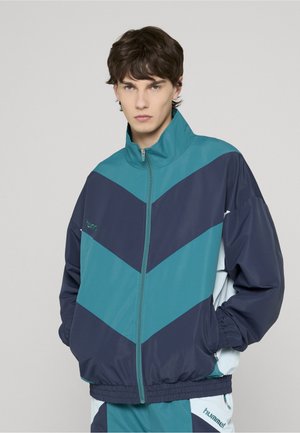 Giacca con zip in blu scuro e verde acqua con motivo a chevron. Realizzata in tessuto liscio, presenta polsini elasticizzati e collo alla coreana. Logo del marchio sul petto.