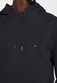 Sudadera de algodón azul marino con capucha ajustable, detalles en metal plateado y un pequeño logotipo bordado en rojo, blanco y azul en el pecho.