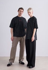 Sorte oversized t-shirts med farverige logo-mønstre, parret med løse beige og sorte bukser, båret af to modeller mod en neutral baggrund.