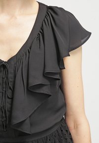 Blusa nera con volant sovrapposti sulle maniche e un dettaglio con lacci sulla parte anteriore, realizzata in tessuto leggero con una consistenza liscia.