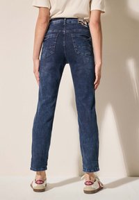 Jean taille haute bleu foncé avec une coupe classique, cinq poches et une légère texture délavée. Associés à des baskets beiges avec des accents rouges.