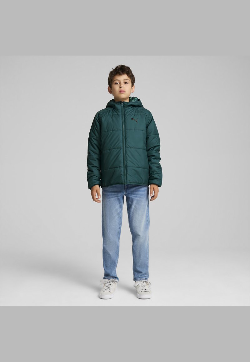 Chaqueta acolchada verde con capucha, con cierre de cremallera y diseño acolchado. Combinada con vaqueros azules y zapatillas blancas.