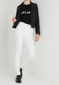 Chaqueta de motociclista de cuero negro, camiseta negra ajustada con texto blanco, jeans blancos y zapatillas negras; diseño elegante y colores contrastantes.
