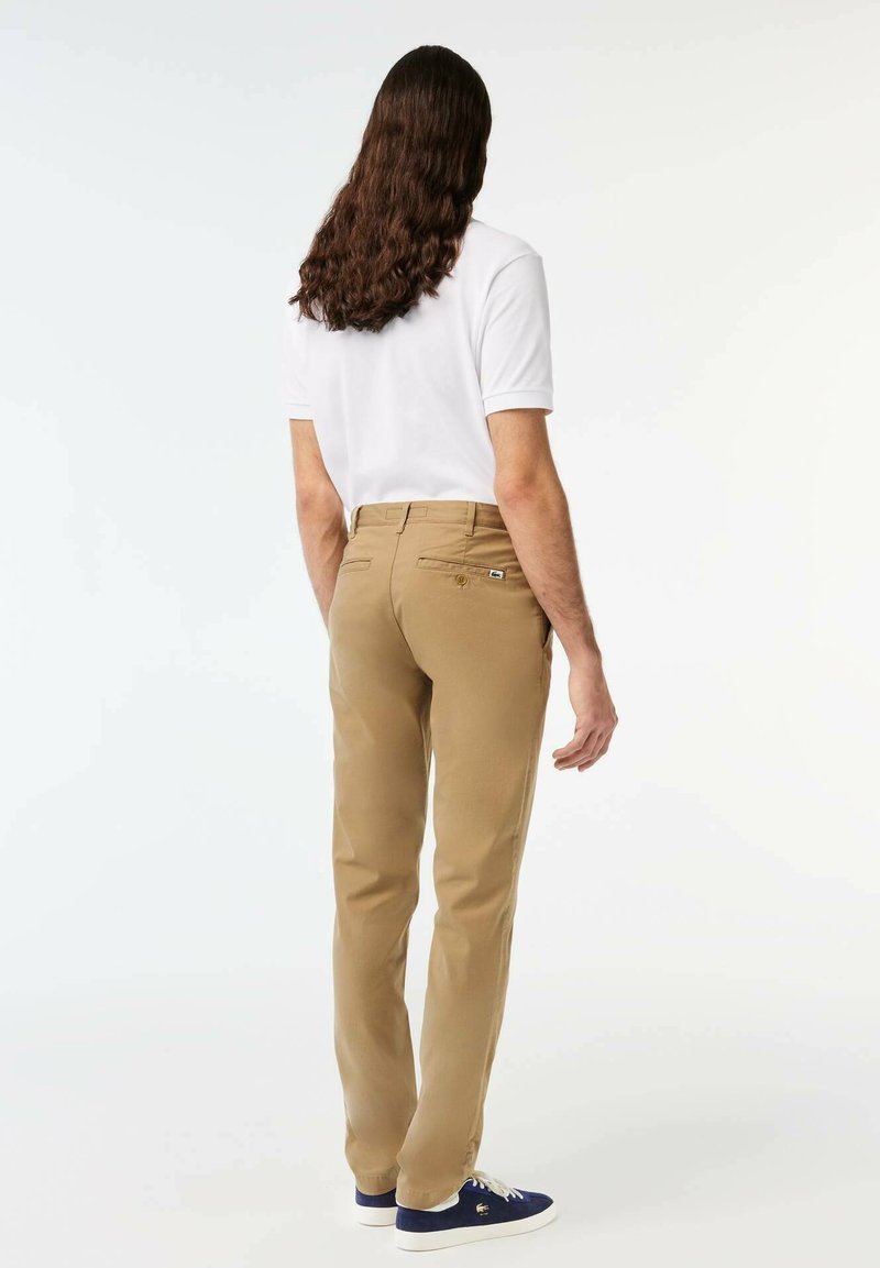 Mann mit langen braunen Haaren, der ein weißes kurzärmliges Polohemd, beige Hosen und marineblaue Sneakers trägt und mit dem Rücken zur Kamera vor einem weißen Hintergrund steht.