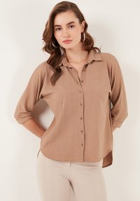 Light brown button-up shirt with a textured fabric, rolled sleeves, and side slits. Features a classic collar and button details.