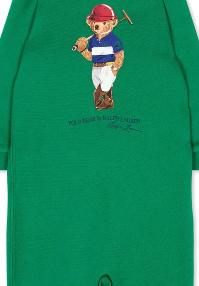 Tessuto verde con un orso che indossa un casco da polo rosso, una maglia a righe blu, pantaloni bianchi, stivali marroni, che tiene una mazza da polo sulla spalla.