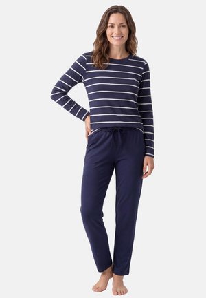 Donna sorridente che indossa una maglia a maniche lunghe a righe blu navy e bianco e pantaloni blu navy a tinta unita con coulisse, in piedi a piedi nudi su uno sfondo semplice.