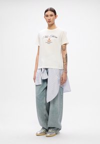 White cotton t-shirt with graphic print; layered striped long shirt tied at waist; loose, faded blue denim jeans; silver shoes with tan soles.