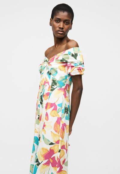 Γυναίκα με κοντά μαλλιά, ντυμένη με λευκό φόρεμα έξωμη με πολύχρωμη ανθισμένη εκτύπωση, στέκεται μπροστά σε απλό ανοιχτό φόντο.