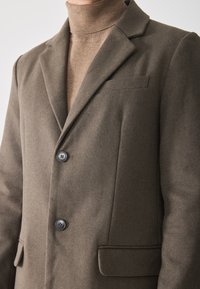 Hombre con un blazer de lana marrón sobre un suéter de cuello alto beige, abotonado en la parte delantera, que muestra las solapas y los bolsillos.