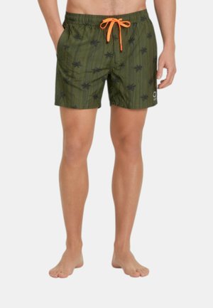 Uomo con pantaloncini da bagno verdi con stampe di palme nere e coulisse arancione, in piedi a piedi nudi su uno sfondo semplice.