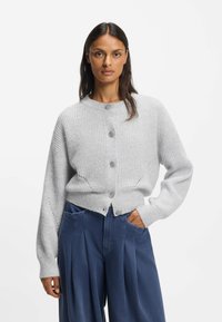 Harmaa ribbineulottu neuletakki, jossa on pyöreä pääntie, etunapit ja laskoshihat, yhdistetty leveisiin sinisiin housuihin ja etutaskuihin.