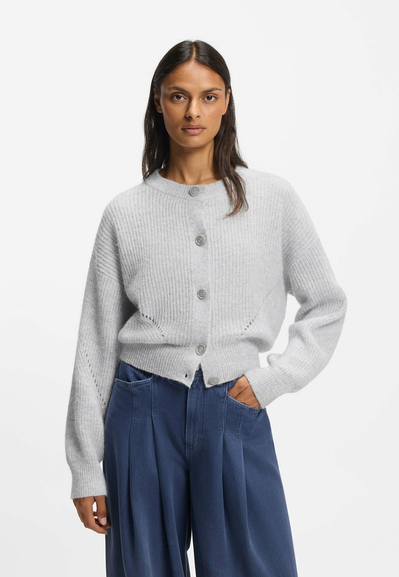 Harmaa ribbineulottu neuletakki, jossa on pyöreä pääntie, etunapit ja laskoshihat, yhdistetty leveisiin sinisiin housuihin ja etutaskuihin.