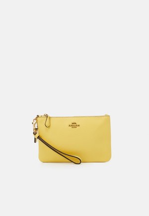 Pochette bracelet jaune Coach avec logo doré et sangle de poignet bordée de noir sur fond blanc.