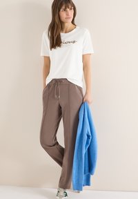 Femme portant une chemise blanche à manches courtes et un pantalon marron à cordon de serrage, tenant un pull bleu, debout devant un fond beige clair.