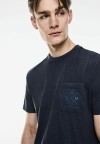 Námornícké bavlněné tričko s krátkým rukávem a kulatým výstřihem. Na levé straně hrudníku je kapsa s modrým potiskem a grafickým designem.