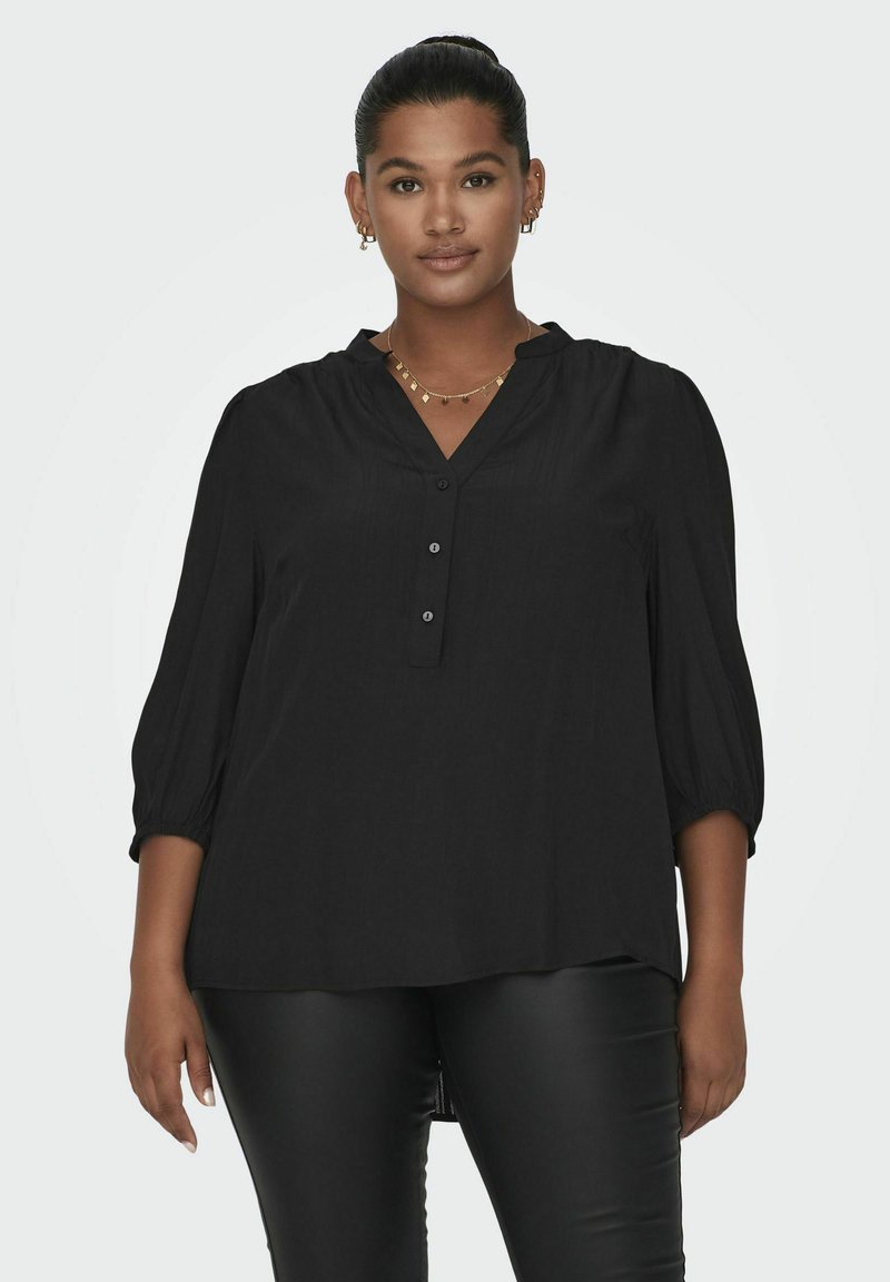 Blusa negra con escote en V, detalle de botones y mangas abullonadas de tres cuartos. Textura suave, corte relajado y líneas de costura sutiles.
