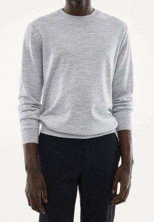 Homme portant un pull gris clair à manches longues et un pantalon noir, debout devant un fond blanc uni.