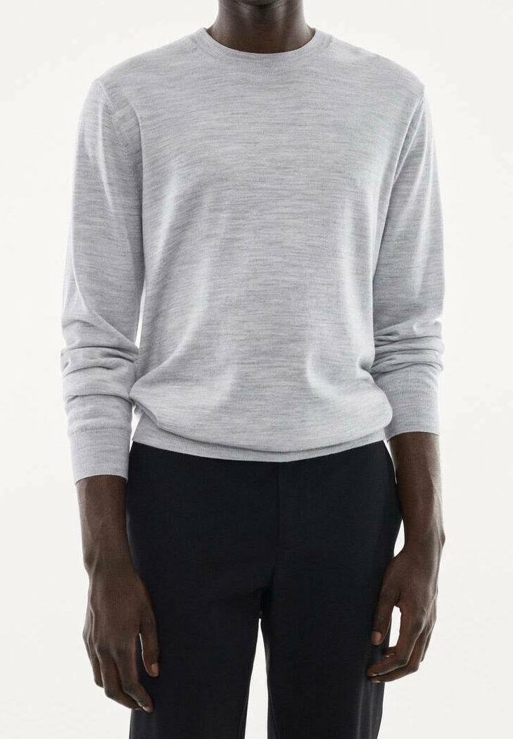 Homme portant un pull gris clair à manches longues et un pantalon noir, debout devant un fond blanc uni.