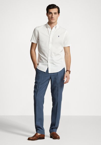Chemise blanche à manches courtes avec un logo sur la poitrine gauche, associée à un pantalon bleu sur mesure et des chaussures habillées marron. Texture de tissu lisse.