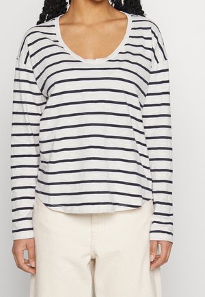 Person iført en hvid langærmet top med marineblå horisontale striber og beige bukser, vist fra hals til hofter mod en ensfarvet baggrund.