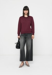 Sudadera marrón con el texto en relieve "PARÍS", combinada con jeans sueltos grises, tacones de punta gris y un bolso negro texturizado.