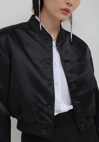Czarna satynowa kurtka typu bomber z zapięciami na guziki, ściągaczami na mankietach i zaokrąglonym kołnierzem; noszona na białej koszuli zapinanej na guziki.