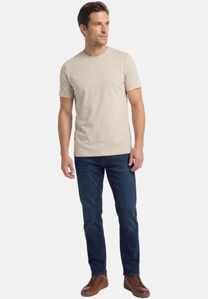 Mężczyzna stojący, ubrany w beżowy, krótki rękaw, niebieskie dżinsy i brązowe skórzane trampki, patrzący lekko w prawo.