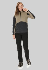 Veste en polaire verte et noire avec fermeture éclair à l'avant, deux poches latérales et manches longues. Associée à un pantalon noir et des chaussures de randonnée.